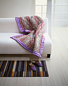Gestreifte Quilt-Tagesdecke auf schlicht weißem Sofa, davor lila Hausschuhe auf gestreiftem Läufer