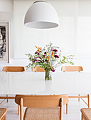 White lamp above vase of flowers on white dining table with wooden chairs