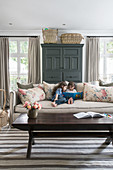 Two children reading on sofa with scatter cushions, dark cupboard flanked by windows and wooden coffee table in foreground