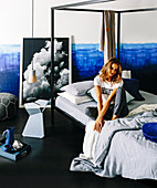 Young woman sitting on the four-poster bed in front of a two-tone wall