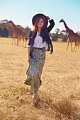 A brunette woman wearing a hat, a white shirt-blouse, a long skirt and a jacket with giraffes in the background