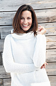 A brunette woman wearing a white knitted jumper standing in front of a wooden wall