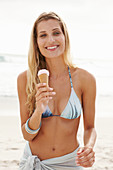 Reife blonde Frau mit Eis im Bikini und Strandtuch am Strand