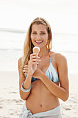Reife blonde Frau mit Eis im Bikini und Strandtuch am Strand