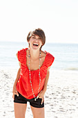 A mature brunette woman on a beach wearing a red shirt, a necklace and black shorts