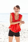 Reife brünette Frau mit rotem Shirt, Halskette und schwarzer Shorts am Strand