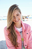 A young blonde woman on a beach wearing a t-shirt and a pink denim jacket