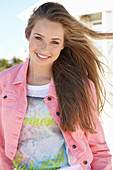 A young blonde woman on a beach wearing a printed t-shirt and a pink denim jacket