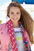 A young blonde woman on a beach wearing a printed t-shirt, a pink denim jacket and a patterned scarf