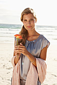 A mature blonde woman on a beach wearing a silver summer dress and holding an orange flower