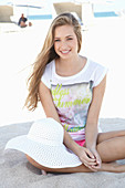 Junge blonde Frau in bedrucktem T-Shirt und mit weißem Hut am Strand