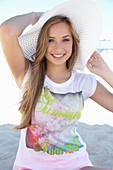 A young blonde woman on a beach wearing a colourful t-shirt and a white summer hat