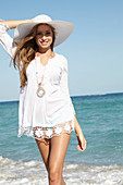 A young blonde woman on a beach wearing a white summer dress, a white summer hat and a pink necklace