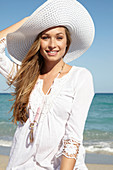 A young blonde woman on a beach wearing a white summer dress, a white summer hat and a pink necklace