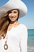 A young blonde woman on a beach wearing a white summer dress, a white summer hat and a pink necklace