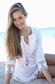 A young blonde woman on a beach wearing a white summer dress and a pink necklace