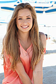 A young blonde woman on a beach wearing a salmon-coloured, off-the-shoulder top