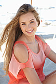 A young blonde woman on a beach wearing a salmon-coloured, off-the-shoulder top