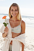 A mature blonde woman on a beach wearing lingerie and a cardigan and holding a flower