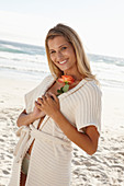 Reife blonde Frau in Dessous und Strickjacke und mit Blume in der Hand am Strand