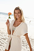 Reife blonde Frau in Dessous und Strickjacke und mit Blume in der Hand am Strand