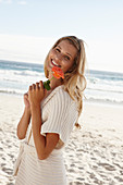 A mature blonde woman on a beach wearing lingerie and a cardigan and holding a flower