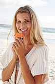 A mature blonde woman on a beach wearing lingerie and a cardigan and holding a flower