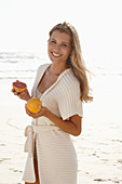 Reife blonde Frau mit Grapefruit in Dessous und Strickjacke am Strand