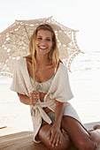 Reife blonde Frau in Dessous und Strickjacke mit Sonnenschirm am Strand