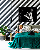Large-format photography on a black and white striped wall, blonde woman lying on bed with green day cover