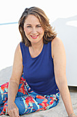 A brunette woman on a beach wearing a purple top and colourful trousers