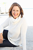 A brunette woman on a beach wearing a white knitted jumper and dark trousers