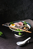 Traditional Japanese Noodle Soup with shiitake mushroom, egg, sliced beef and greens in ceramic bowl with wooden chopsticks