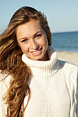 A young blonde woman on a beach wearing a white turtle neck jumper