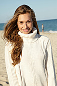 A young blonde woman on a beach wearing a white turtle neck jumper