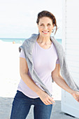 Brunette woman wearing lilac T-shirt, jeans and grey sweater over shoulders on beach
