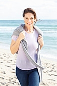 Brünette Frau in lila T-Shirt, mit grauem Pulli über den Schultern und Jeans am Strand