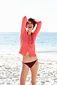 A mature brunette woman on a beach wearing a black bikini and a red shirt