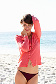 A mature brunette woman on a beach wearing a black bikini and a red shirt