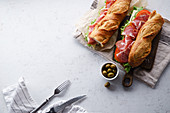 Top view of two fresh baguette sandwiches bahn-mi styled with olives, ham, sliced cheese, tomatoes and fresh lettuce