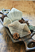 Prism-shaped Christmas decorations made from folded book pages and pine cones in wooden dish