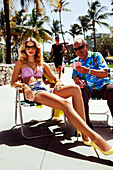 A young blonde woman wearing a bikini sitting on a chair and playing cards with an older man
