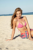 A young blonde woman by the sea wearing a pink bikini with a beach towel around her hips