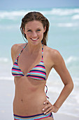 A young brunette woman on a beach wearing a striped bikini