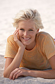 A mature blonde woman with short hair on a beach wearing a striped bikini and an orange top