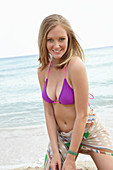 A young blonde woman on a beach wearing a purple bikini