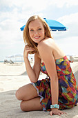 A young blonde woman on a beach wearing a colourful summer dress