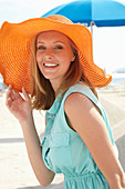 Junge blonde Frau im hellblauen Sommerkleid und orangenem Sommerhut am Strand