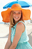 A young blonde woman on a beach wearing a light-blue summer dress and an orange summer hat