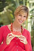 Reife Frau mit kurzen blonden Haaren im roten Shirt und einem Stück Wassermelone in der Natur